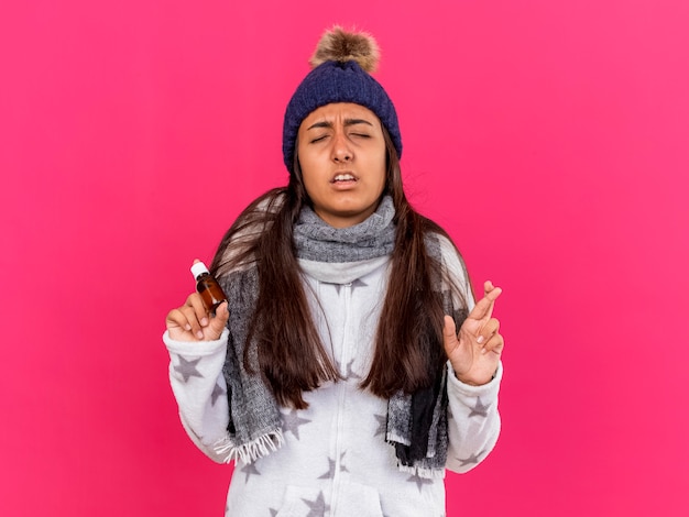 Free photo concerned young ill girl with closed eyes wearing winter hat with scarf holding medicine in glass bottle crossing fingers isolated on pink