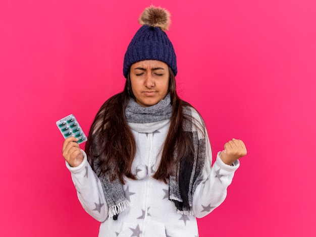 Concerned young ill girl with closed eyes wearing winter hat with scarf holding pills isolated on pink background