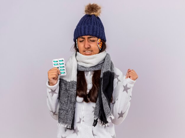 Concerned young ill girl with closed eyes wearing winter hat with scarf holding pills putting pills under hat and showing yes gesture isolated on white background