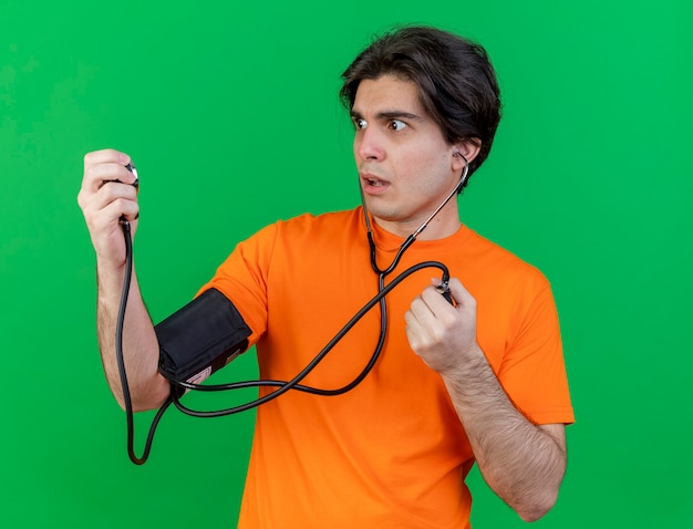 Concerned young ill man measuring his own pressure with sphygmomanometer isolated on green