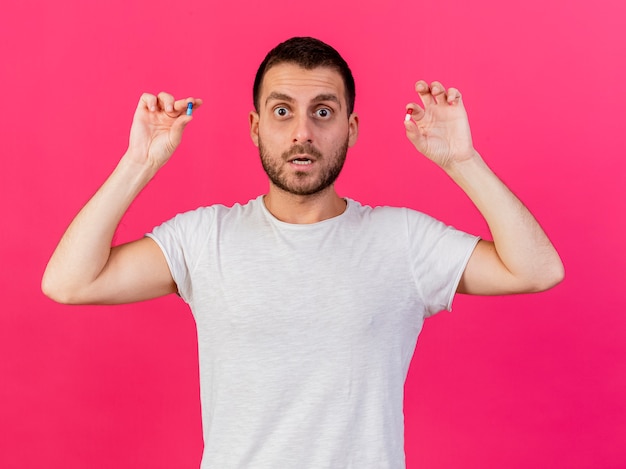 Concerned young ill man raising pills isolated on pink background