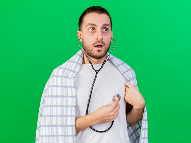 Concerned young ill man wearing and listen to his own heartbeat with stethoscope isolated on green background