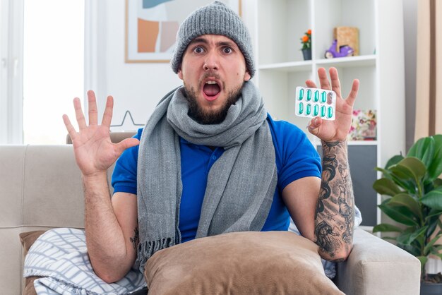 concerned young ill man wearing scarf and winter hat sitting on sofa in living room with pillow on his legs showing pack of capsules and empty hand looking at front with open mouth
