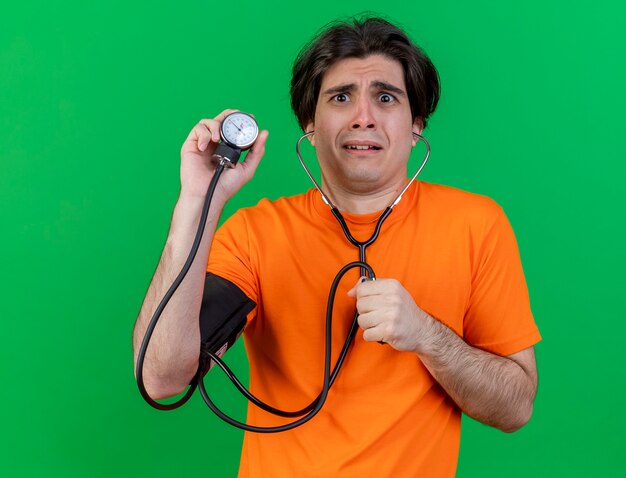 緑に分離された血圧計を保持している聴診器を身に着けている心配している若い病気の人