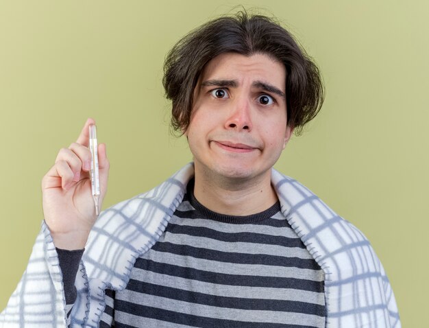 Concerned young ill man wrapped in plaid holding thermometer isolated on olive green