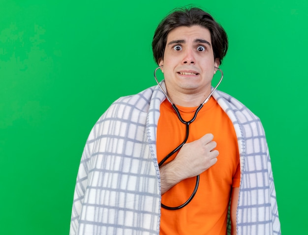 Concerned young ill man wrapped in plaid wearing and listening to his own heartbeat with stethoscope isolated on green background