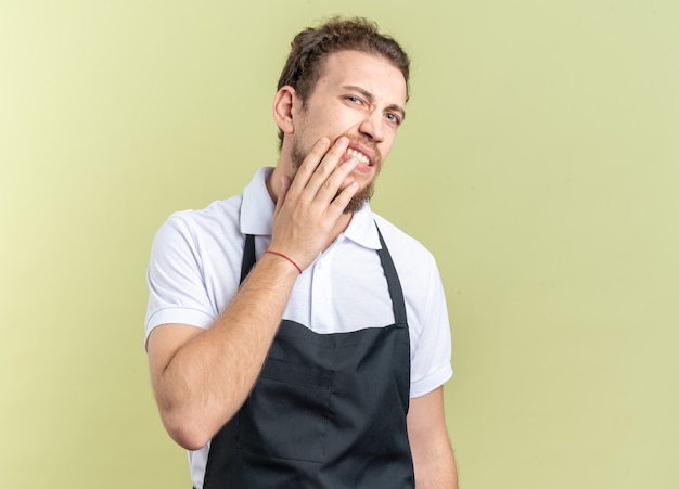 Concerned young male barber wearing uniform covered mouth with hand isolated on olive green wall