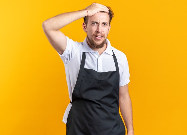 Concerned young male barber wearing uniform putting hand on forehead isolated on yellow wall