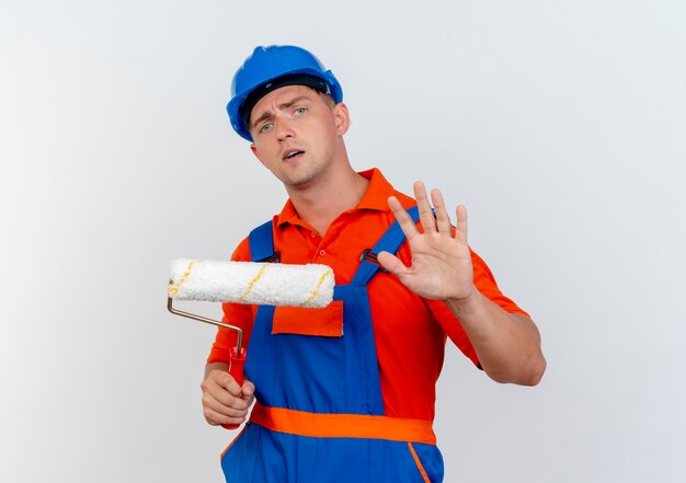 Foto gratuita giovane costruttore maschio interessato che indossa l'uniforme e il casco di sicurezza che tiene il rullo di vernice e che mostra il gesto di arresto