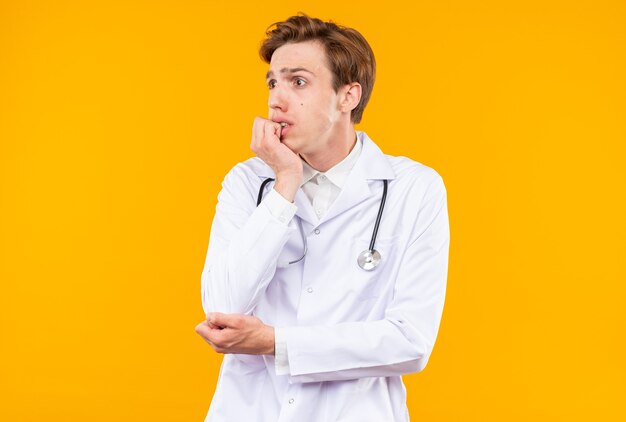 Concerned young male doctor wearing medical robe with stethoscope bites nails isolated on orange wall
