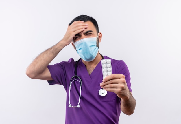 紫色の外科医の服と聴診器の医療用マスクを身に着けている心配している若い男性医師は、孤立した白い壁の額に手を置きます