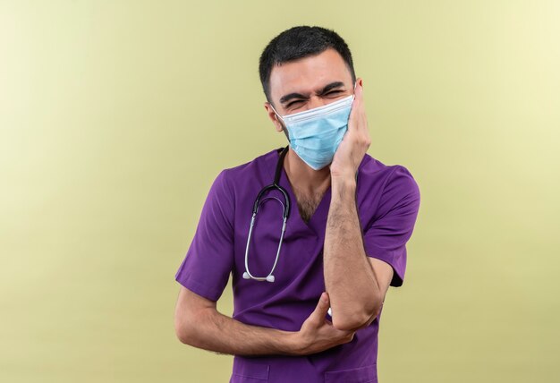 Обеспокоенный молодой мужчина-врач в фиолетовой хирургической одежде и медицинской маске со стетоскопом положил руку на щеку на изолированной зеленой стене