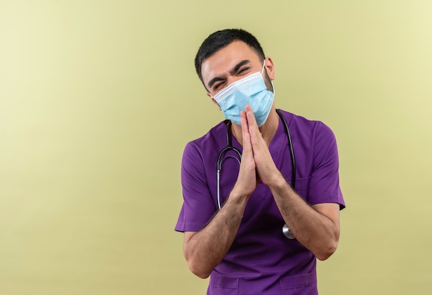紫色の外科医の服と聴診器の医療マスクを身に着けている心配している若い男性医師は、孤立した緑の壁に祈りのジェスチャーを示しています