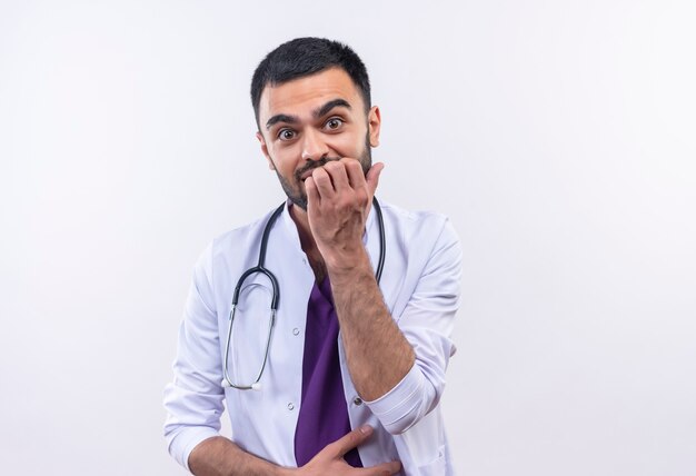 Concerned young male doctor wearing stethoscope medical gown put his hand on chin on isolated white wall