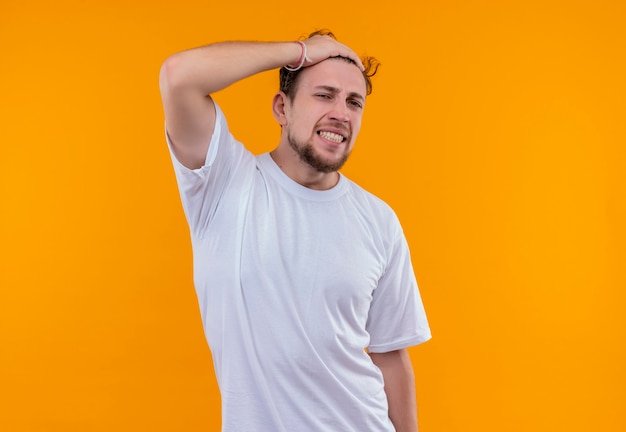 Concerned young man wearing white t-shirt put his hand on head on isolated orange wall