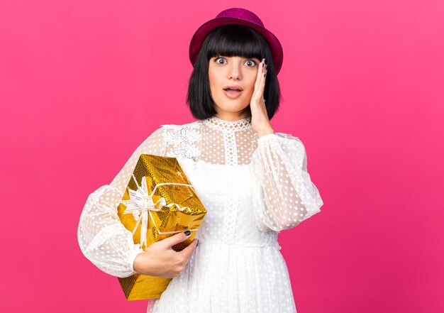 Concerned young party girl wearing party hat holding gift package keeping hand on face looking at front isolated on pink wall with copy space