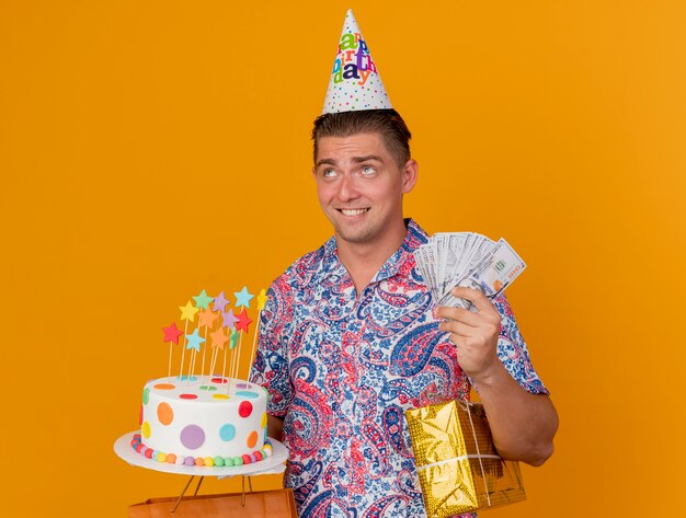 Concerned young party guy looking at up wearing birthday cap holding cake with gifts and cash isolated on orange