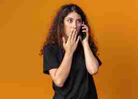 Free photo concerned young pretty girl talking on phone keeping hand on mouth looking at side isolated on orange wall with copy space