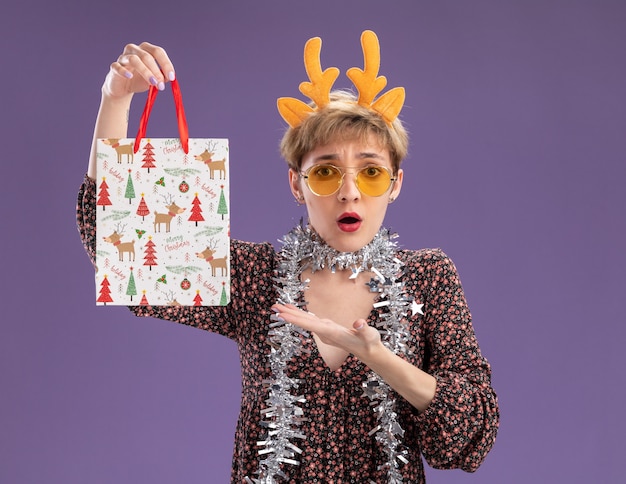 Concerned young pretty girl wearing reindeer antlers headband and tinsel garland around neck with glasses holding and pointing at christmas gift bag looking at camera isolated on purple background