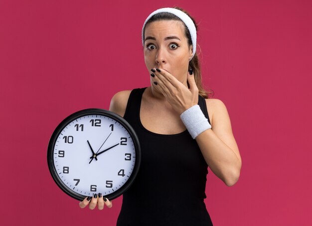 concerned young pretty sporty woman wearing headband and wristbands holding clock keeping hand on mouth