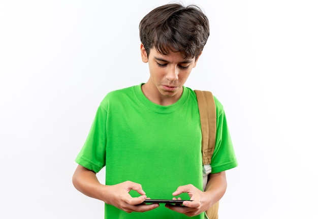 Concerned young school boy wearing backpack play on phone isolated on white wall