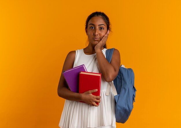 Concerned young schoolgirl wearing backpack holding books and putting hand on cheek 