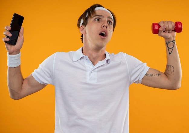 Concerned young sporty guy wearing headband and wristband raising phone with dumbbell isolated on orange wall