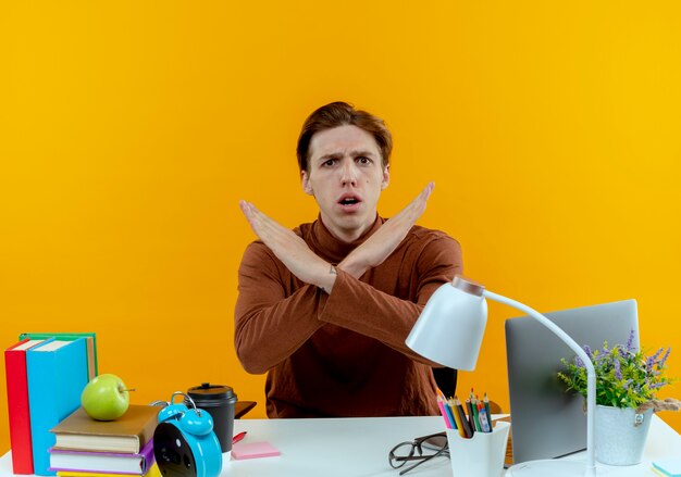 Concerned young student boy sitting at desk with school tools showing gesture of no 