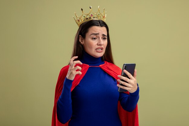 Concerned young superhero girl wearing crown holding and looking at phone isolated on olive green