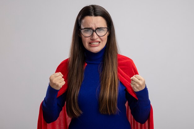 Concerned young superhero girl wearing glasses holding fists isolated on white background