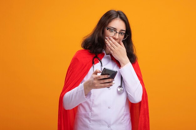 Обеспокоенная молодая женщина-супергерой в униформе доктора и стетоскоп в очках, держащая и смотрящая на мобильный телефон, держа руку за рот, изолированную на стене с копией пространства