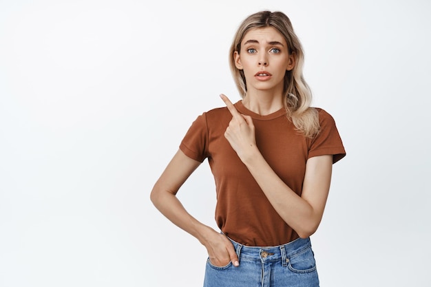 Concerned young woman pointing finger at upper left corner looking worried at camera standing in brown casual tshirt and jeans white background