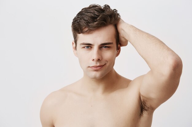 Condifent young european man with stylish haircut and appealing eyes, being naked, touching dark hair, posing . Handsome male model with healthy clean skin gently smiling