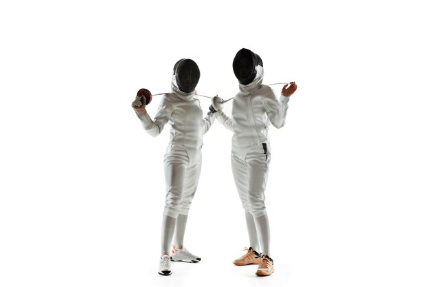 Confidence. Teen girls in fencing costumes with swords in hands on white background. Young female models practicing and training in motion, action. Copyspace.