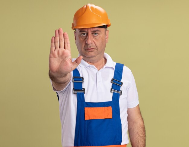 Confident adult builder man gestures stop hand sign isolated on olive green wall