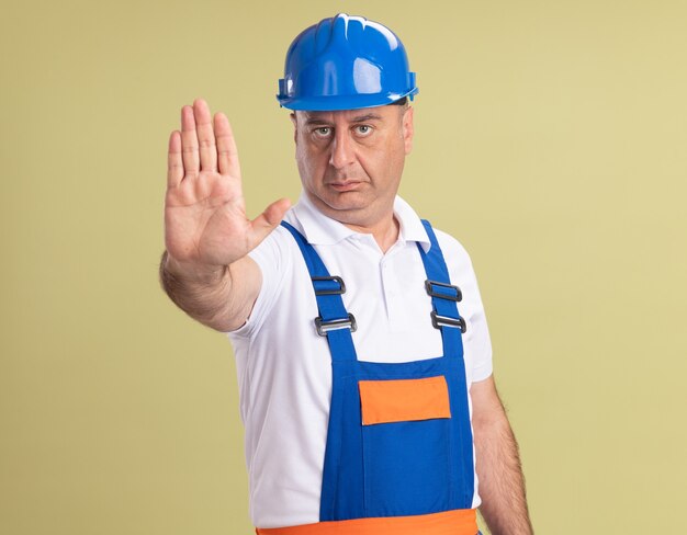 Confident adult builder man in uniform gestures stop hand sign isolated on olive green wall