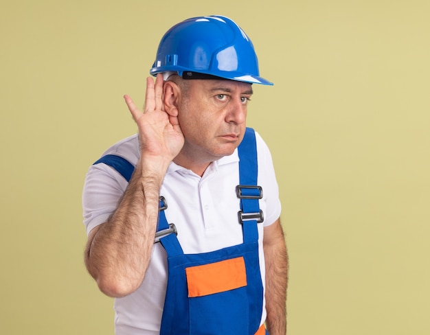 Confident adult builder man in uniform holds hand behind ear isolated on olive green wall