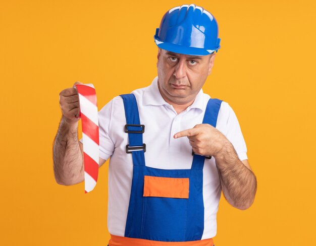 Confident adult builder man in uniform holds and points at scotch tape isolated on orange wall