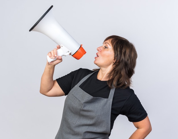 Confident adult caucasian female barber in uniform looking at side and speaking into loud speaker isolated on white background with copy space