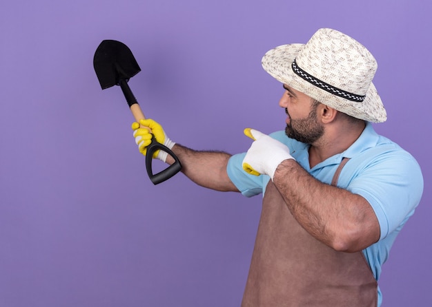 Бесплатное фото Уверенный взрослый кавказский садовник в садовой шляпе и перчатках смотрит и указывает на лопату