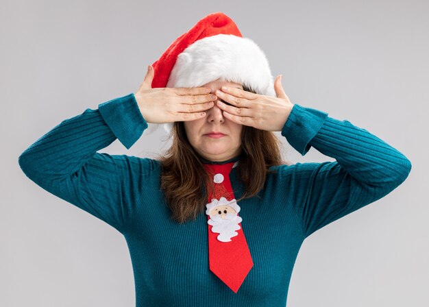 Confident adult caucasian woman with santa hat and santa tie covering eyes with hands isolated on white background with copy space