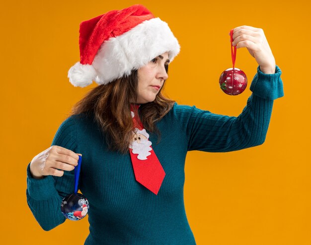 Confident adult caucasian woman with santa hat and santa tie holding and looking at glass ball ornaments isolated on orange background with copy space