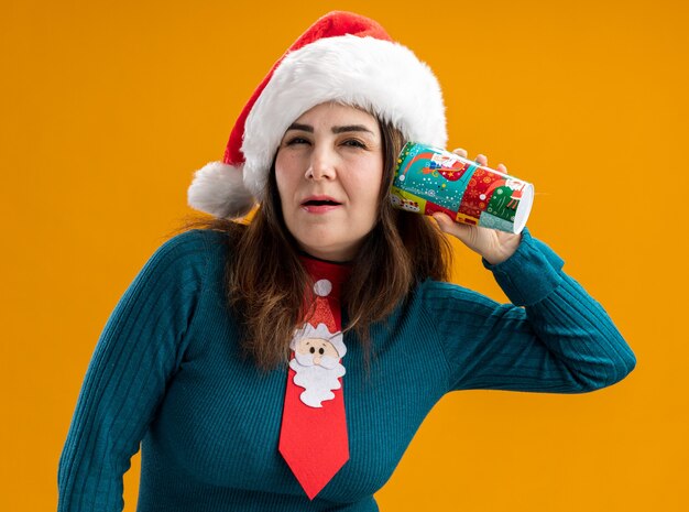 Confident adult caucasian woman with santa hat and santa tie holding paper cup close to ear isolated on orange background with copy space