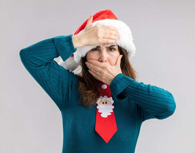 Confident adult caucasian woman with santa hat and santa tie puts hand on forehead and on mouth isolated on white background with copy space