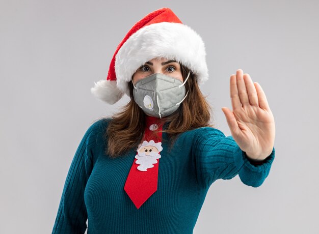 Confident adult caucasian woman with santa hat and santa tie wearing medical mask gesturing stop hand sign 