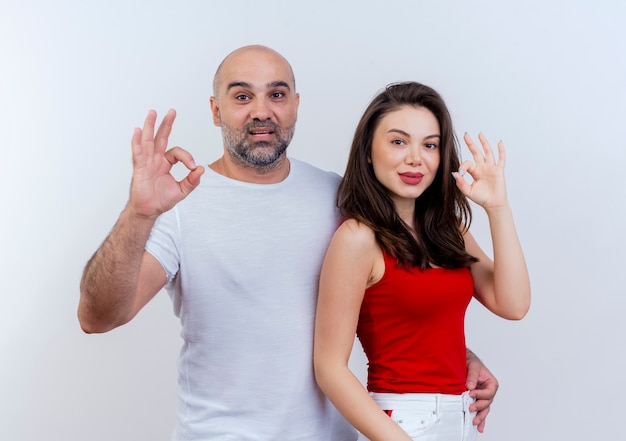 Confident adult couple man holding woman by waist both doing ok signs and  isolated on white wall