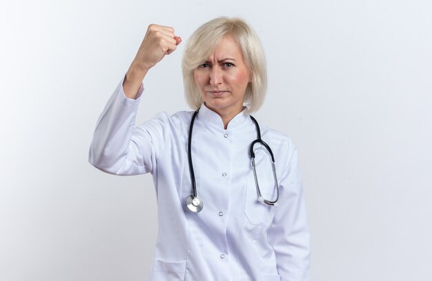 청진기가 있는 의료 가운을 입은 자신감 있는 성인 여성 의사는 복사 공간이 있는 흰 벽에 주먹을 쥐고 있다