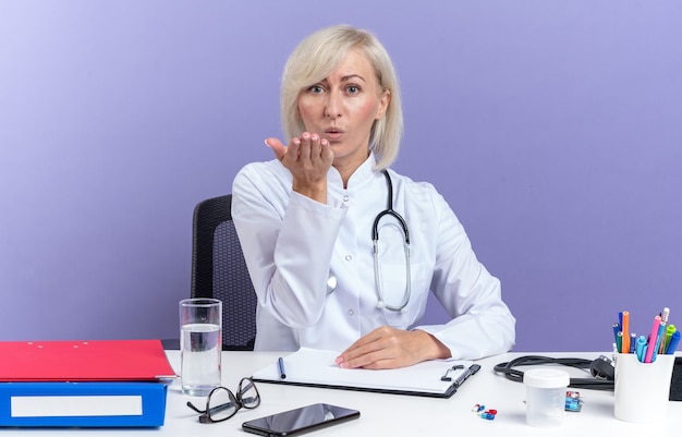 confident adult female doctor in medical robe with stethoscope sitting at desk with office tools sending kiss with hand isolated on purple wall with copy space