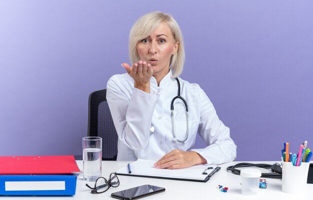 의료 가운을 입은 자신감 있는 성인 여성 의사, 책상에 앉아 있는 청진기, 복사 공간이 있는 보라색 벽에 격리된 손으로 키스를 보내는 사무실 도구