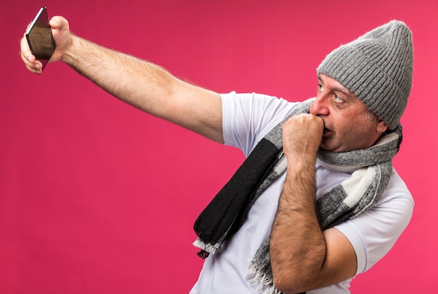 confident adult ill caucasian man with scarf around neck wearing winter hat looking at phone taking selfie and keeping fist close to mouth isolated on pink wall with copy space